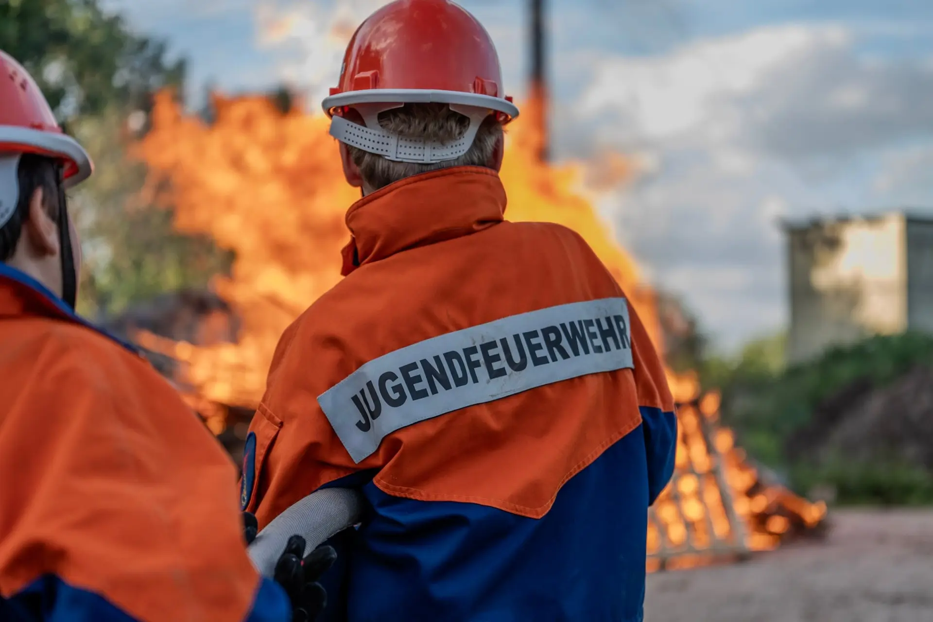 Jugendfeuerwehr vor einem brennenden Holzstapel, von hinten fotografiert
