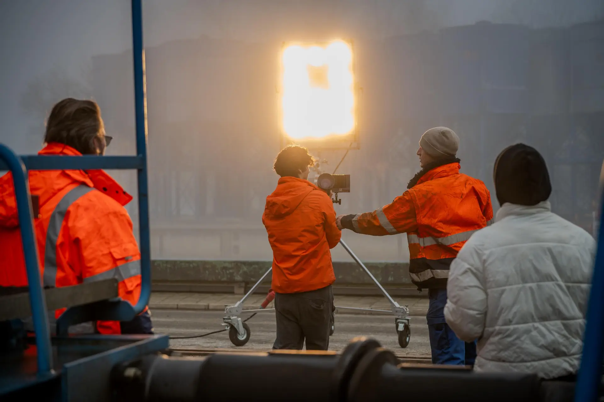 Filmteam in Warnkleidung mit Kamera auf einem Stativwagen vor einer hellen Lichtquelle im Nebel